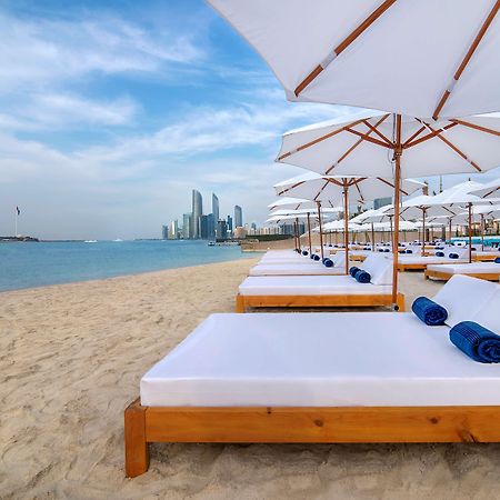 Radisson Blu Hotel & Resort, Abu Dhabi Corniche Екстериор снимка The photo depicts a serene beach setting with several white lounge beds arranged neatly on the sandy shore. Each bed is paired with a white umbrella providing shade. In the background, a skyline of modern buildings can be seen along the coast, hintin