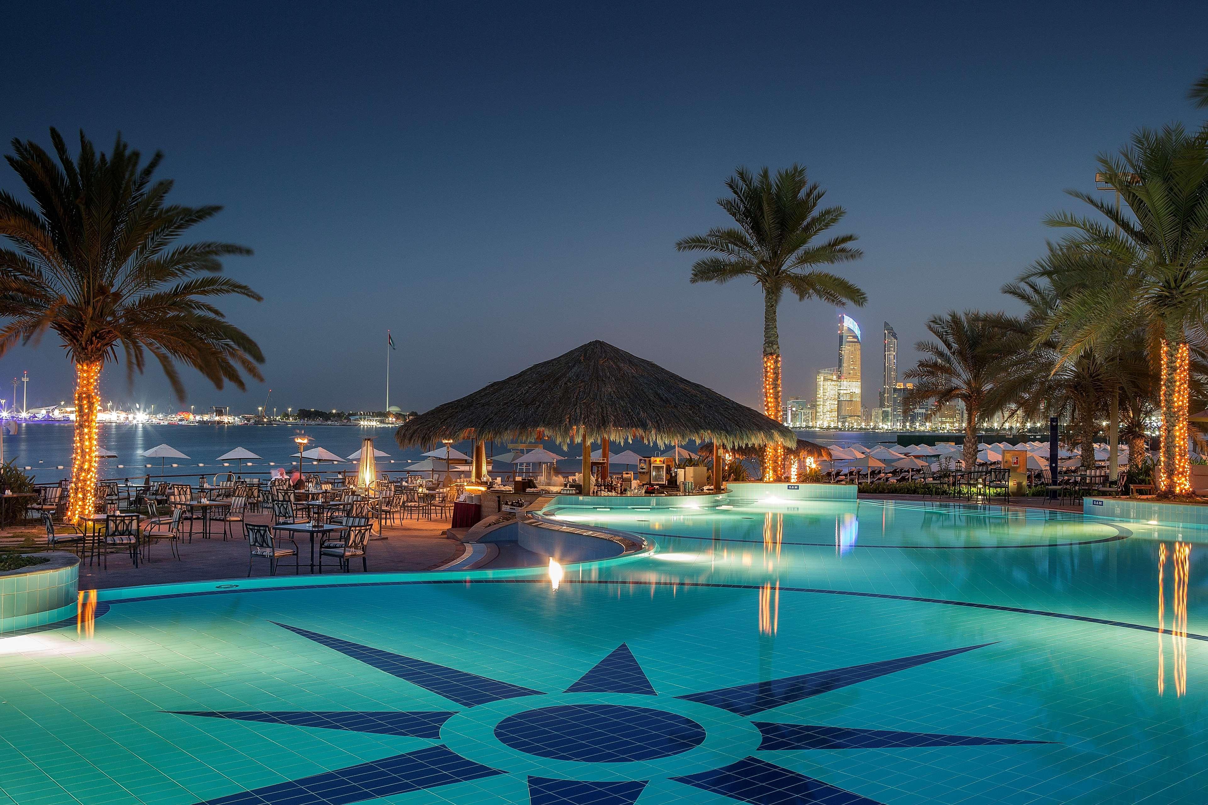Radisson Blu Hotel & Resort, Abu Dhabi Corniche Екстериор снимка The photo shows a picturesque outdoor pool area at night. The pool features a decorative tile pattern resembling a star at its center. Surrounding the pool are tall palm trees, some illuminated by soft lighting. In the background, there is a beachsid