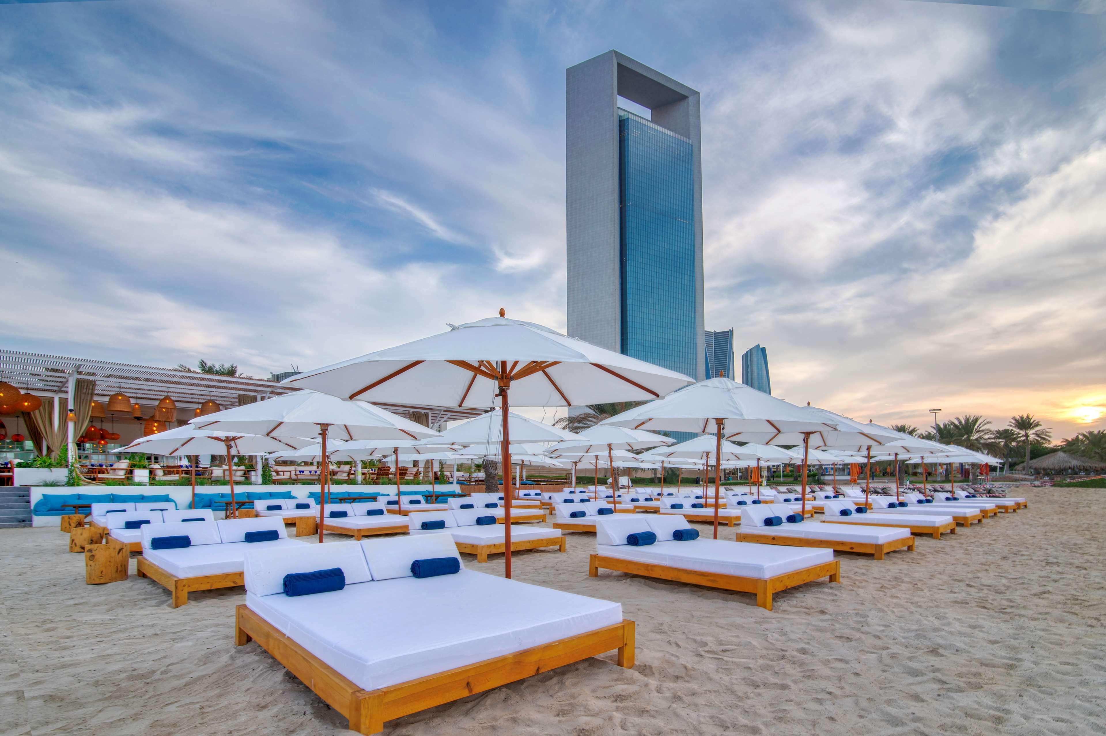 Radisson Blu Hotel & Resort, Abu Dhabi Corniche Екстериор снимка The photo depicts a beach scene with a number of wooden sunbeds arranged on the sandy shore. Each sunbed is equipped with white cushions and blue pillows. Large white umbrellas are positioned above the sunbeds, providing shade. In the background, the