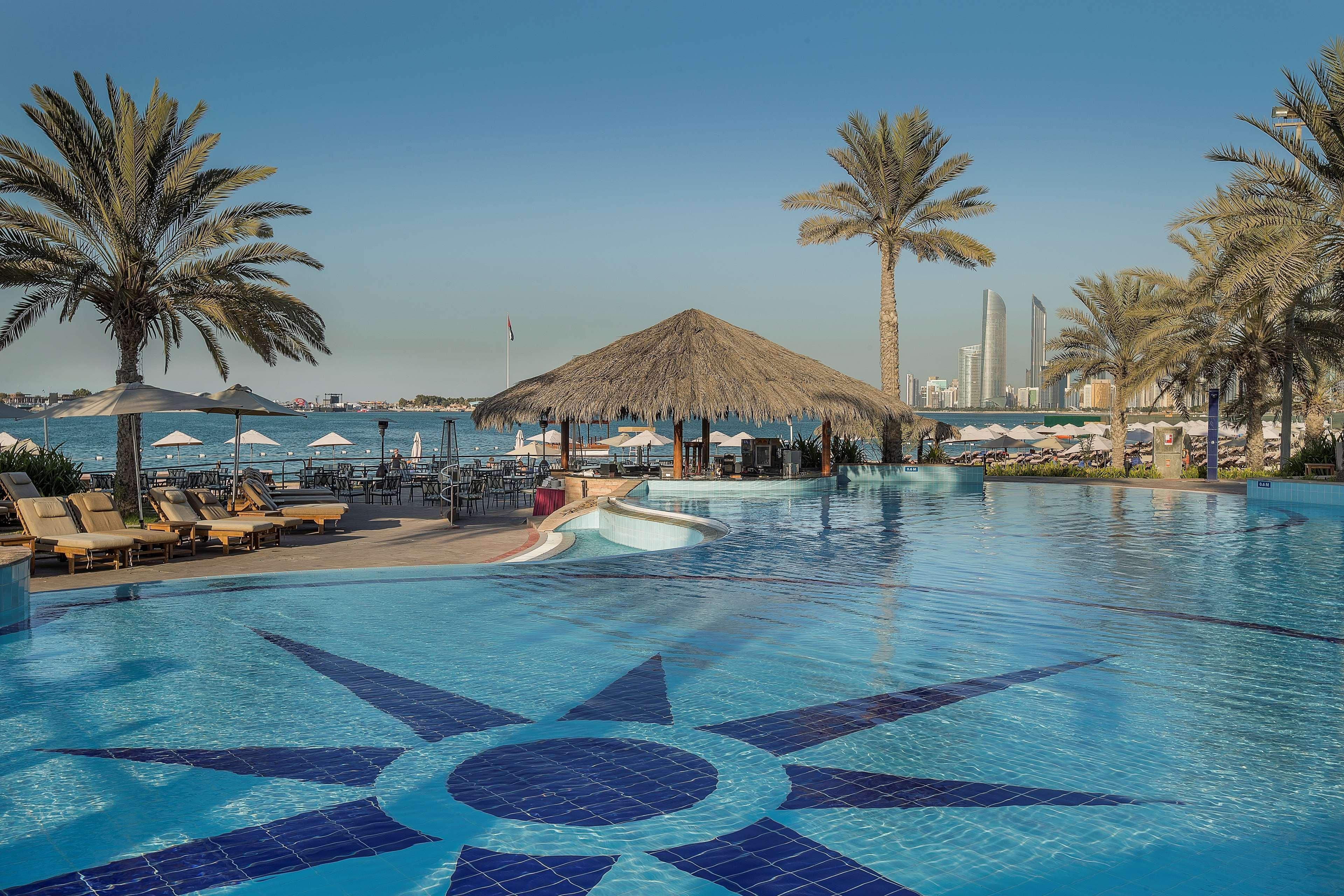 Radisson Blu Hotel & Resort, Abu Dhabi Corniche Екстериор снимка The photo shows a panoramic view of a luxurious resort pool area. In the foreground, there is a large blue swimming pool with a decorative star pattern on the bottom. Surrounding the pool are several lounge chairs, and there are palm trees providing 