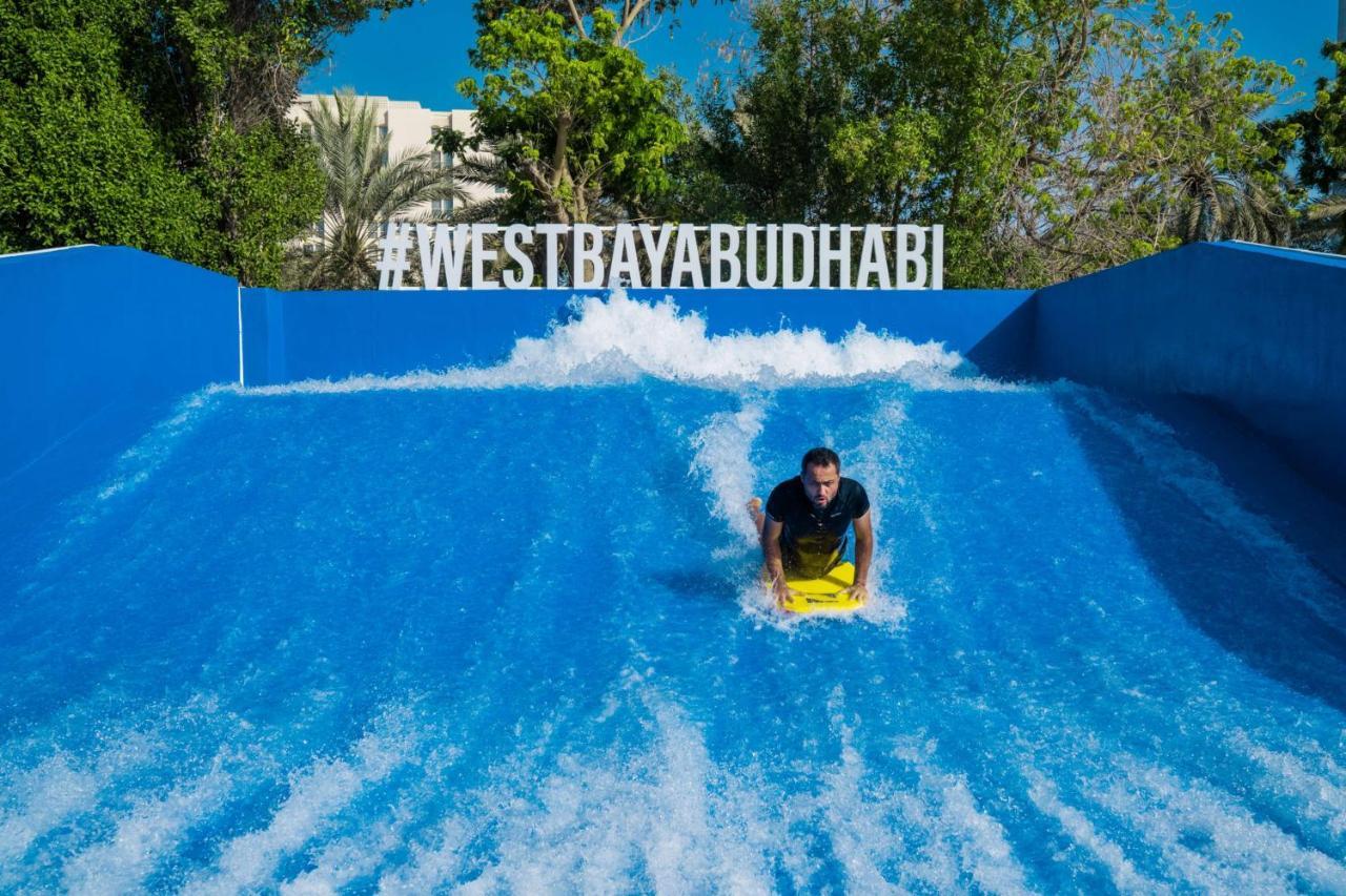 Radisson Blu Hotel & Resort, Abu Dhabi Corniche Екстериор снимка The photo shows a person riding a bodyboard on a wave simulator, creating artificial waves. The background features a bright blue wall with the hashtag #WESTBAYABUDHABI displayed prominently. Surrounding the wave pool, there are trees and greenery, s