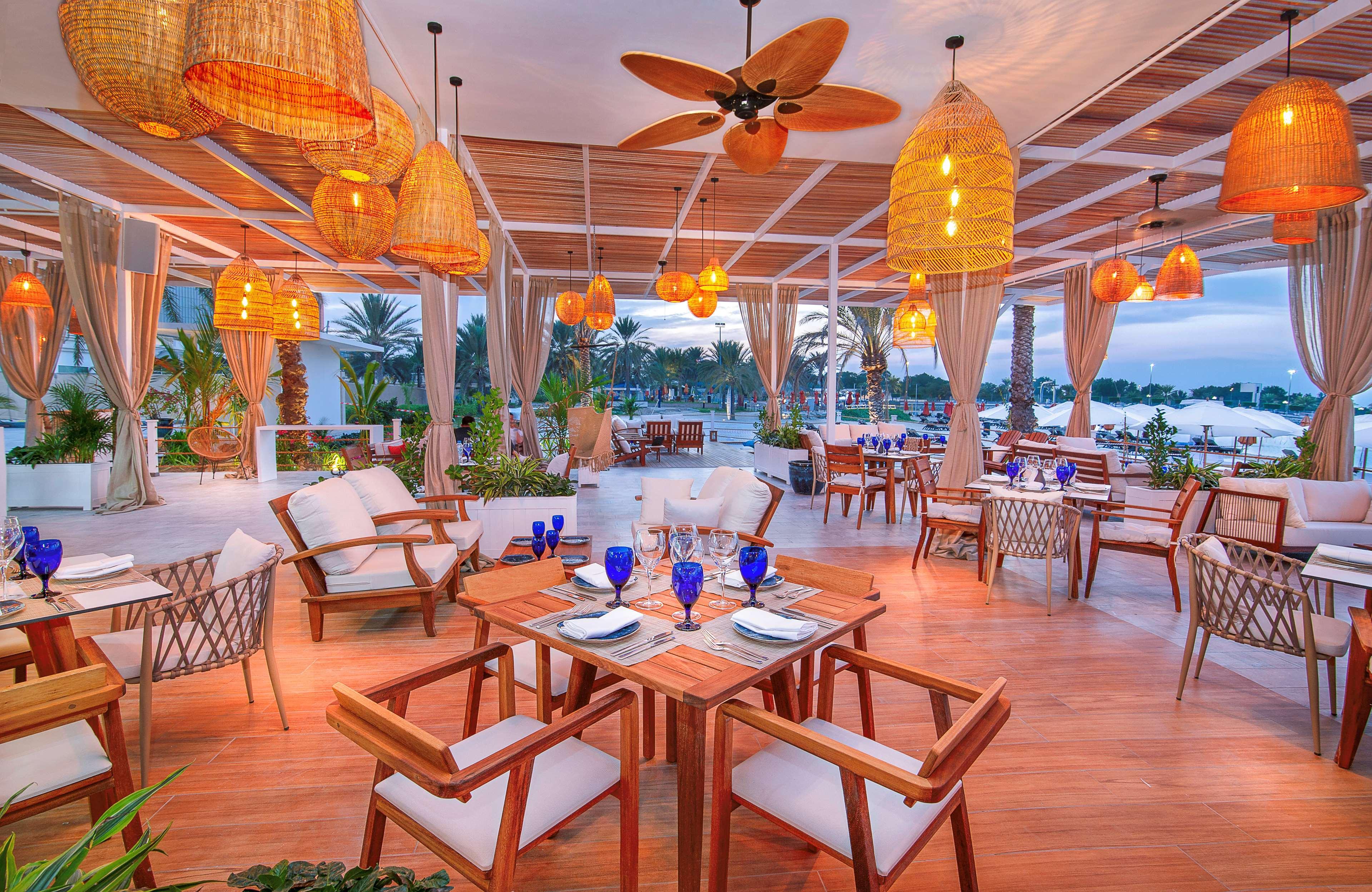 Radisson Blu Hotel & Resort, Abu Dhabi Corniche Екстериор снимка The photo depicts an elegant dining area, likely in a restaurant. The space features wooden tables and comfortable chairs arranged neatly. There are multiple blue glasses set on the tables, suggesting a readiness for dining. The lighting is warm, pro