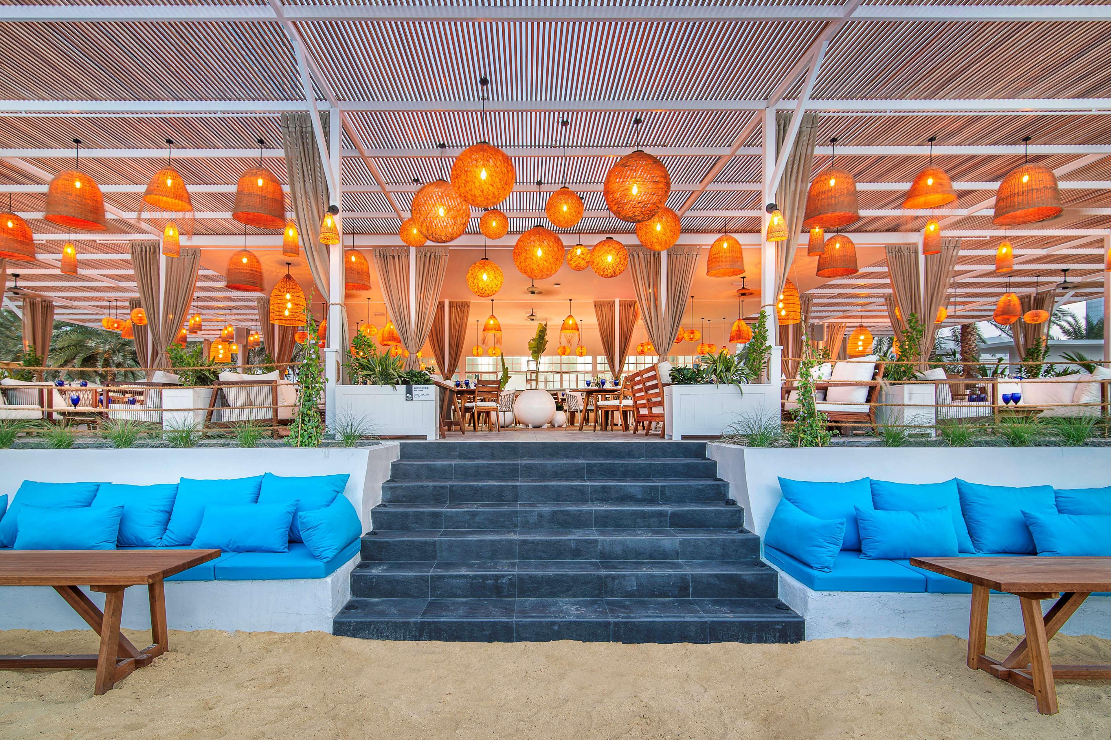 Radisson Blu Hotel & Resort, Abu Dhabi Corniche Екстериор снимка The photo shows a modern, stylish restaurant or lounge area that appears to be located near a beach. The setting features a spacious outdoor dining space with a roof supported by beams. The interior is decorated with warm lighting lamps, which are ha