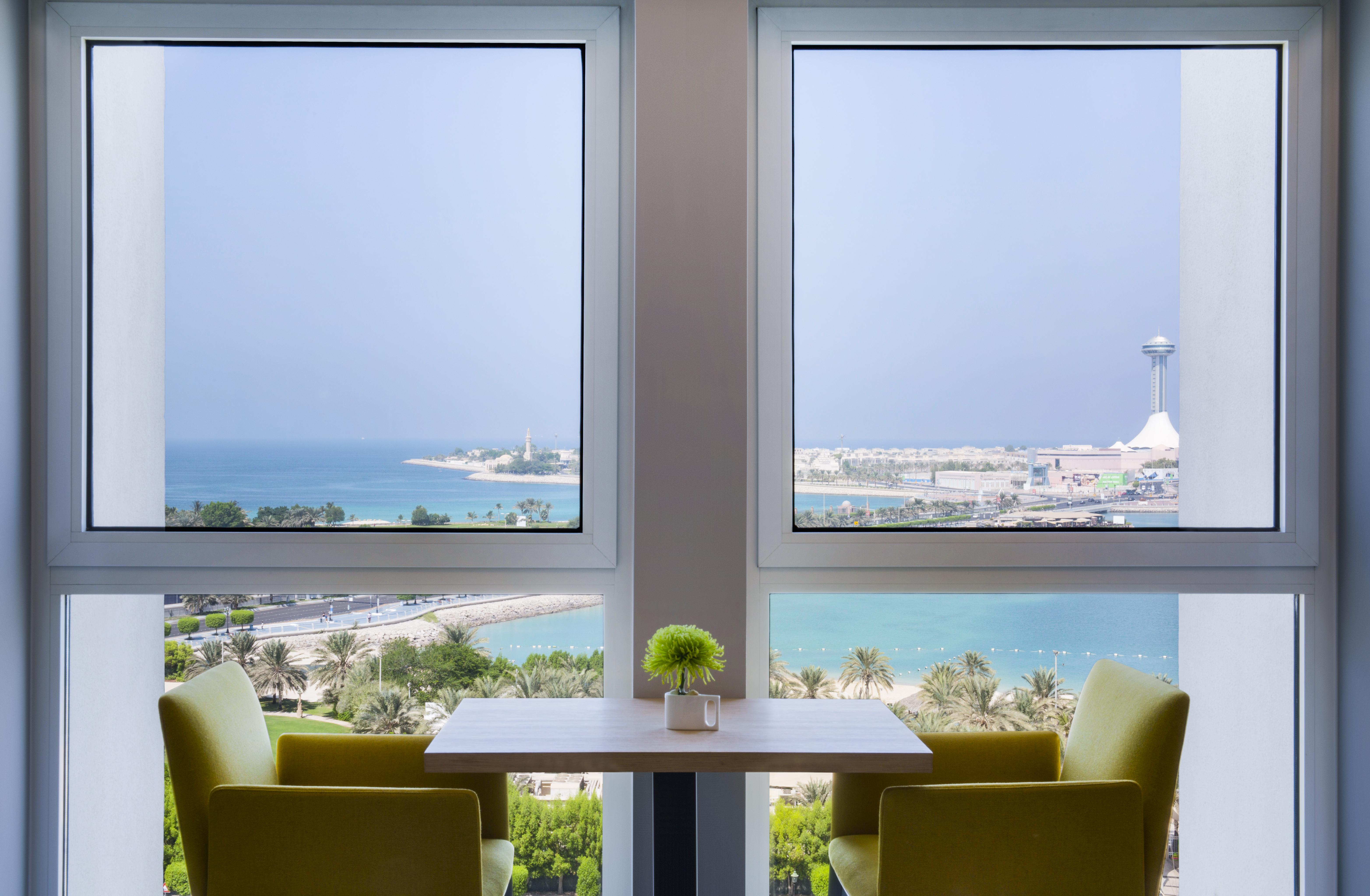 Radisson Blu Hotel & Resort, Abu Dhabi Corniche Екстериор снимка The photo shows a serene interior space featuring two large windows that frame a beautiful view of the ocean and coastline. The scene is bright, with sunlight filtering in, and there are two green chairs positioned around a small table. A small plant